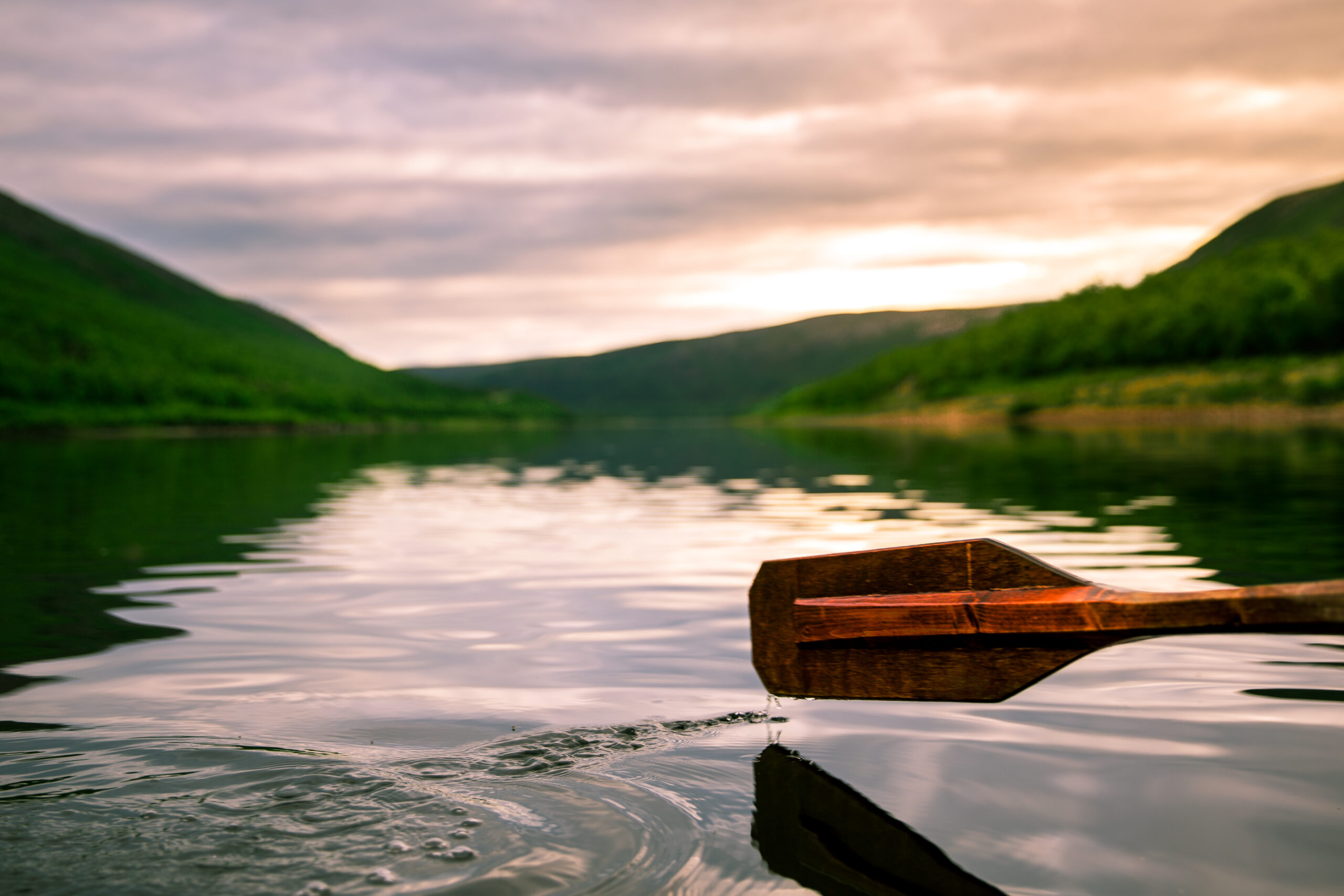A selective focus of a paddle above the surface of a lake with a background of green hills at sunset